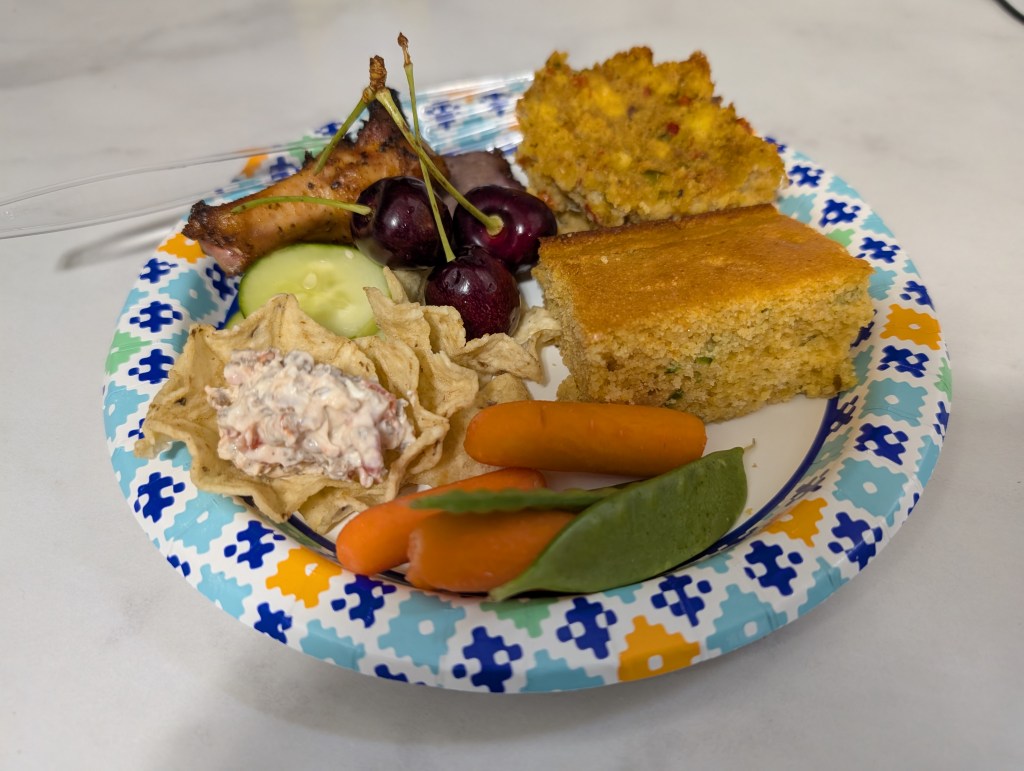 A paper plate loaded with items at a potluck, including a slice of chili cheese cornbread