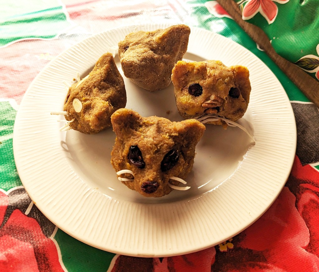 Four molotes de platano formed into little cat heads with eyes and noses made from fruit and nuts, and shredded coconut whiskers