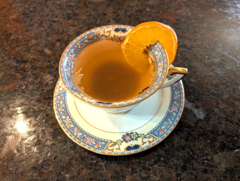 Orange tea in a ceramic teacup with saucer and a sliced mandarin orange on the rim. A few loose leaves float on top.